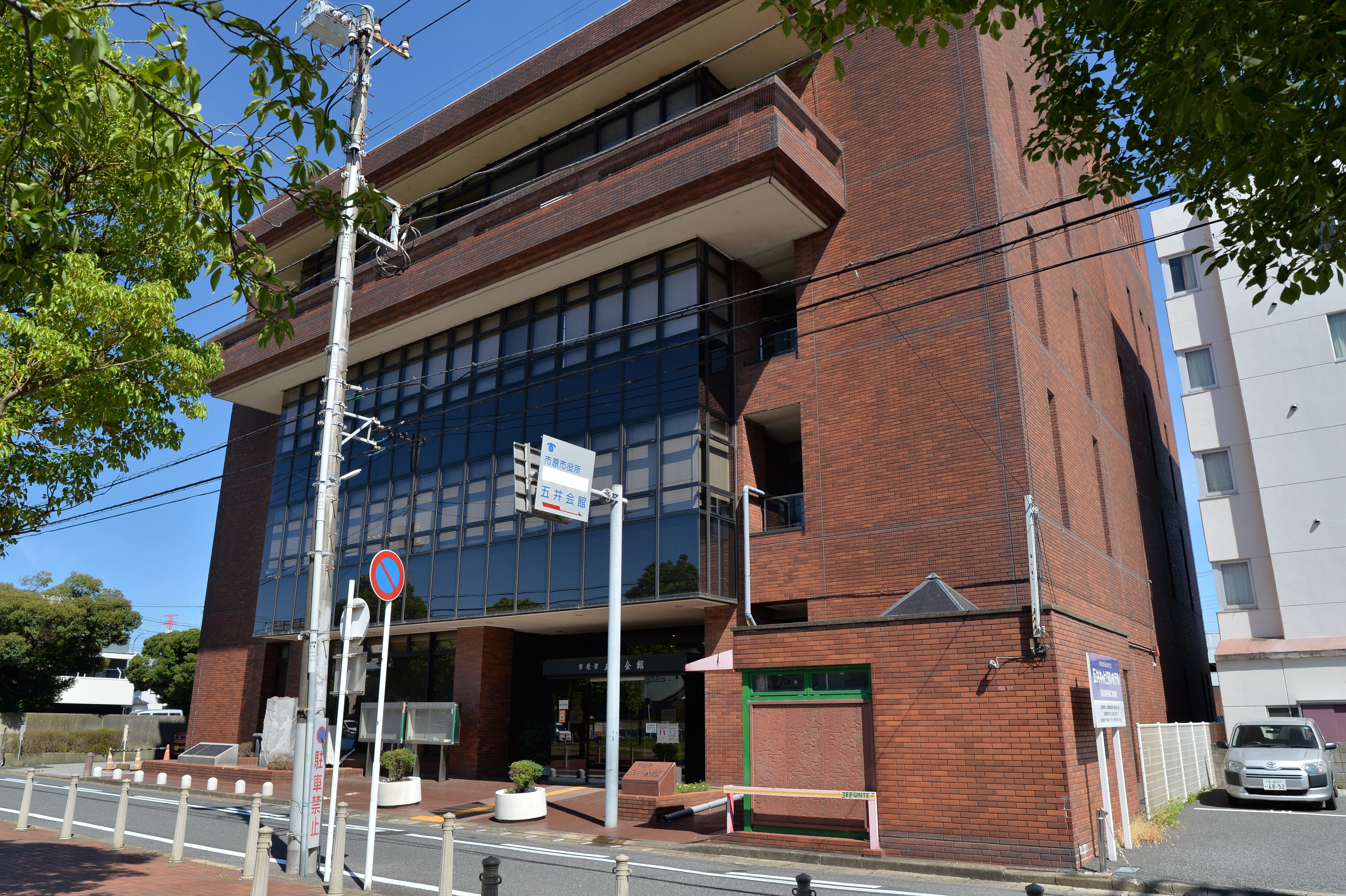 晴天下の斜め前方からの外観全景。赤レンガ外壁とガラスカーテンウォールの4階建。歩道側の看板に「市原市役所 五井会館」表示あり。4928x3280px 2023-07-16撮影
